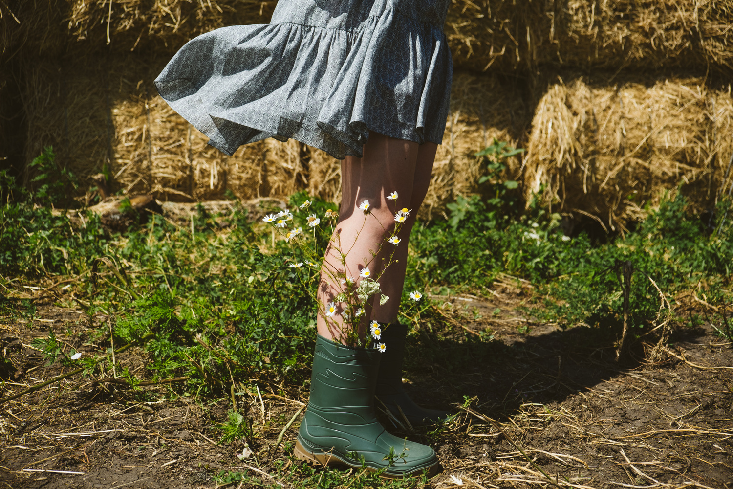 Young girl in rubber boots with flowers standing against the background of straw bales on country farm. Farming, Cottagecore, Farmcore, Countrycore aesthetics, fresh air, countryside, slow life